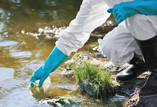 a person in white hazard suit with blue gloves reaches into the water to take a sample
