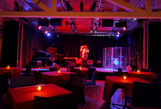 Live band performing on stage in a moody, red-lit music venue with candlelit tables in the foreground