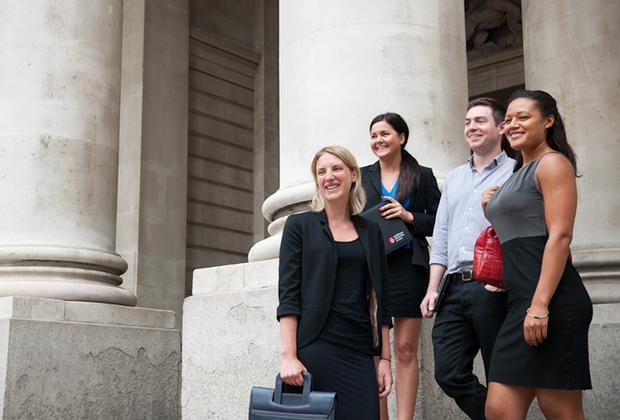 Four professionally dressed students stand outside a building with large stone columns, smiling and holding folders and bags.