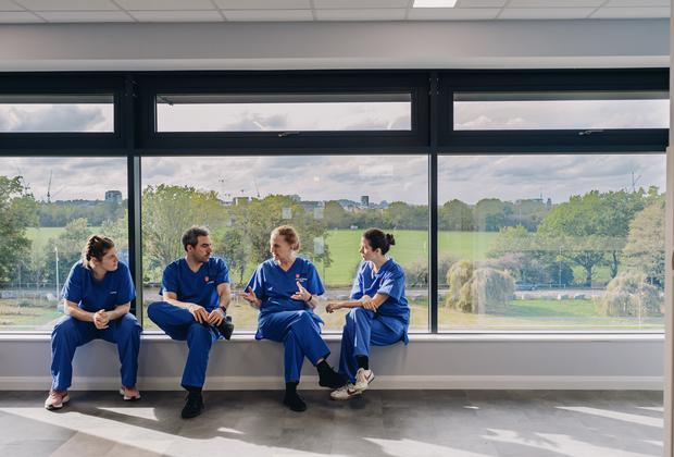 Nursing students in clinical uniforms talk together beside large windows overlooking green space.