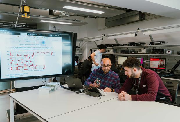 Product design students collaborate with a lecturer on an electronics prototype in a university workshop.