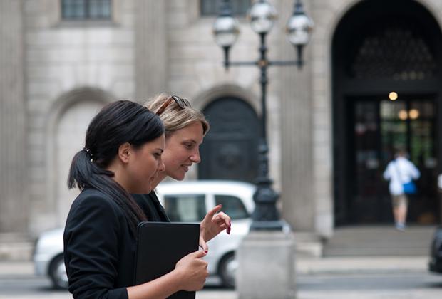 Two students walk and talk outside a university building, carrying study materials.