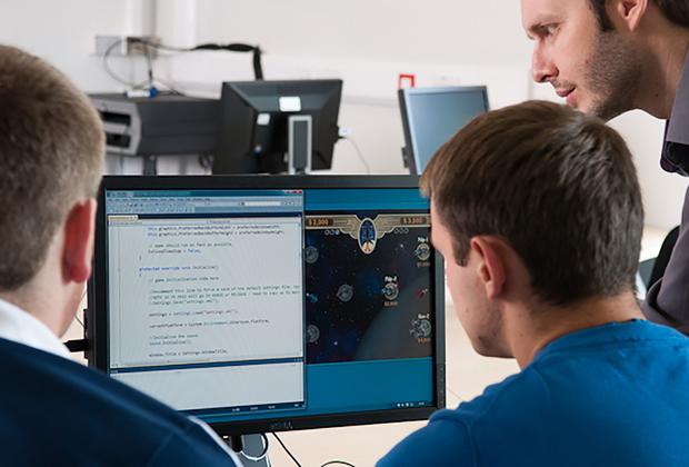 Two men sitting at a desk looking at a computer screen, with a third man standing next to them, leaning over to look at the screen
