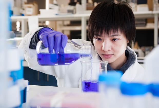 Student in a lab wearing a white coat and blue gloves mixing chemicals