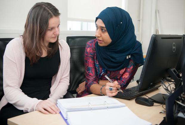Two female students (one wearing a headscarf) sitting at a desk, talking
