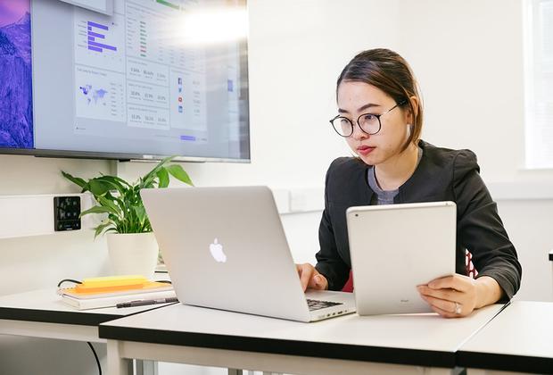 A lady sitting at a desk on her Macbook while holding a bunch of papers in the other hand.