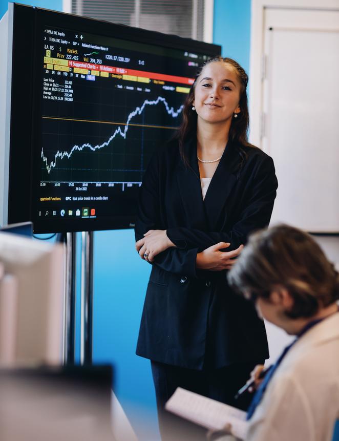 A professional businesswoman stands confidently before a sizable display screen, dressed in a formal business suit.