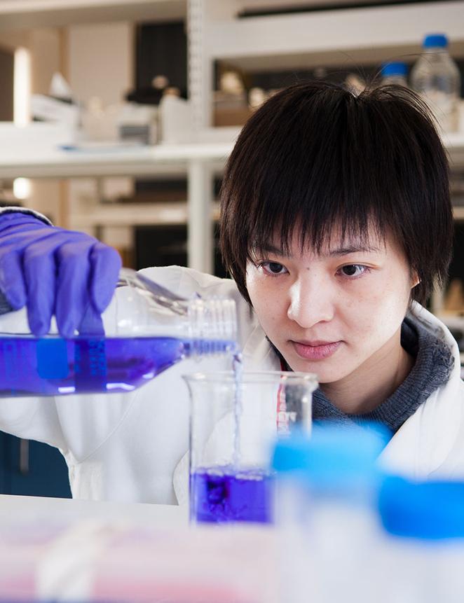 student in lab coat pours purple liquid into beaker