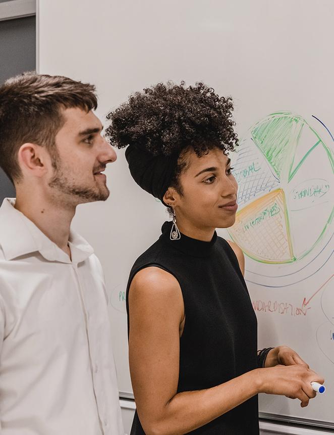 two smartly dressed people stand in front of whiteboard showing pie chart, listening to someone off camera speaking