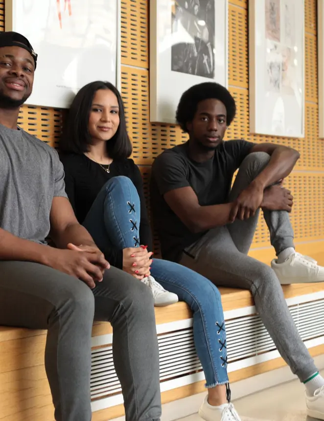 Three young people sitting on a bench in front of a wall, engaged in conversation and enjoying each other's company.
