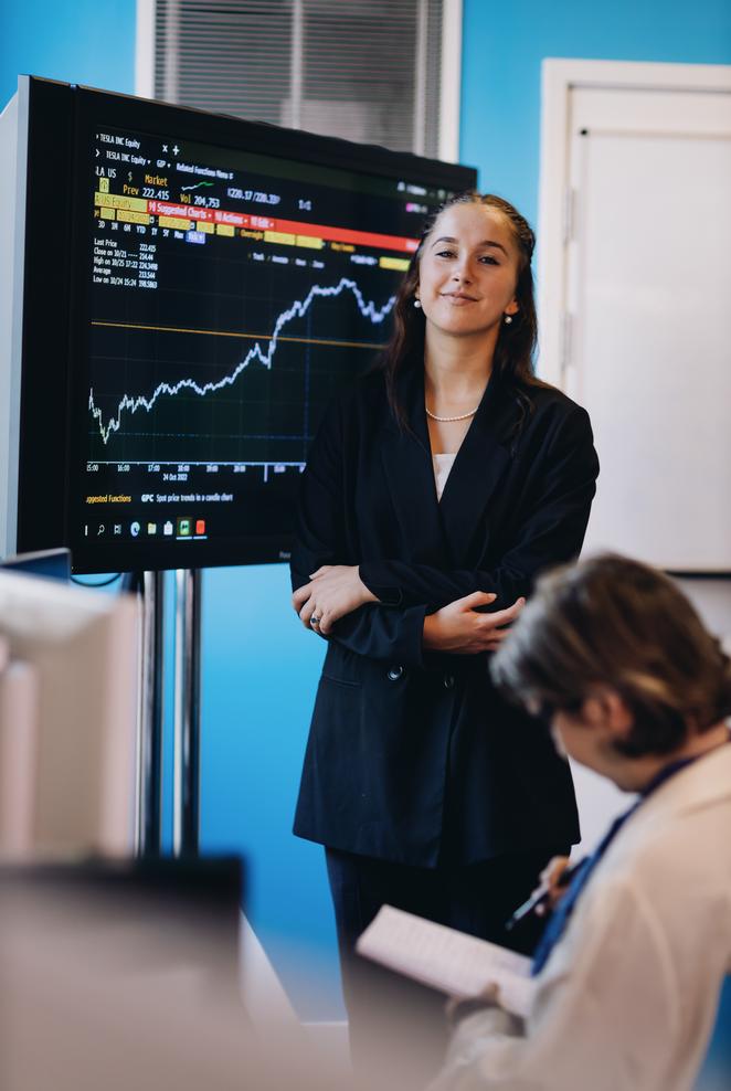 A professional businesswoman stands confidently before a sizable display screen, dressed in a formal business suit.