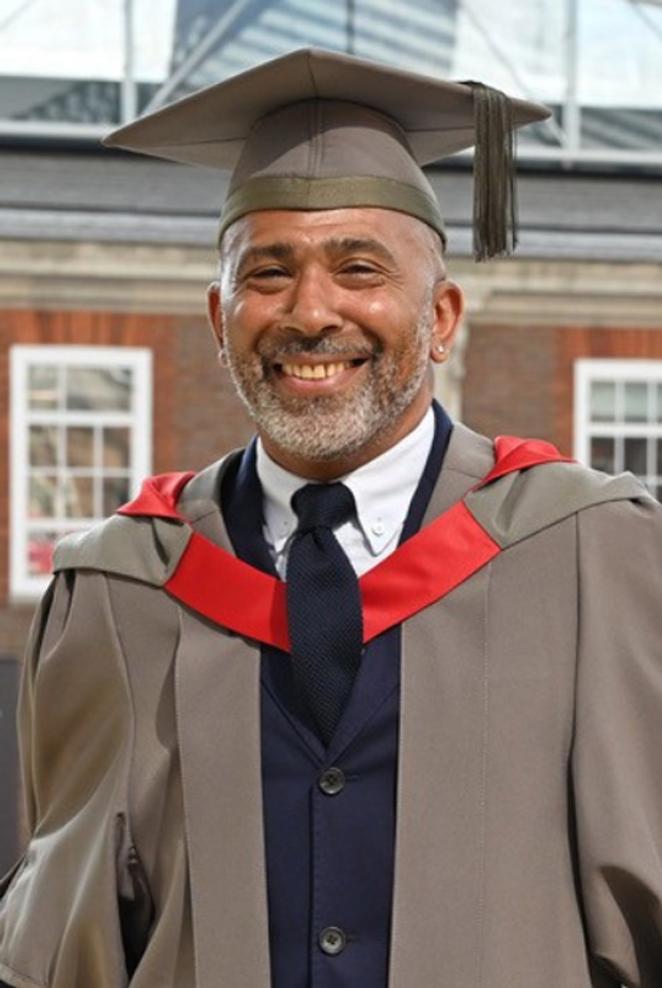 A man in honorary graduates cap and gown poses for a photo in a university building
