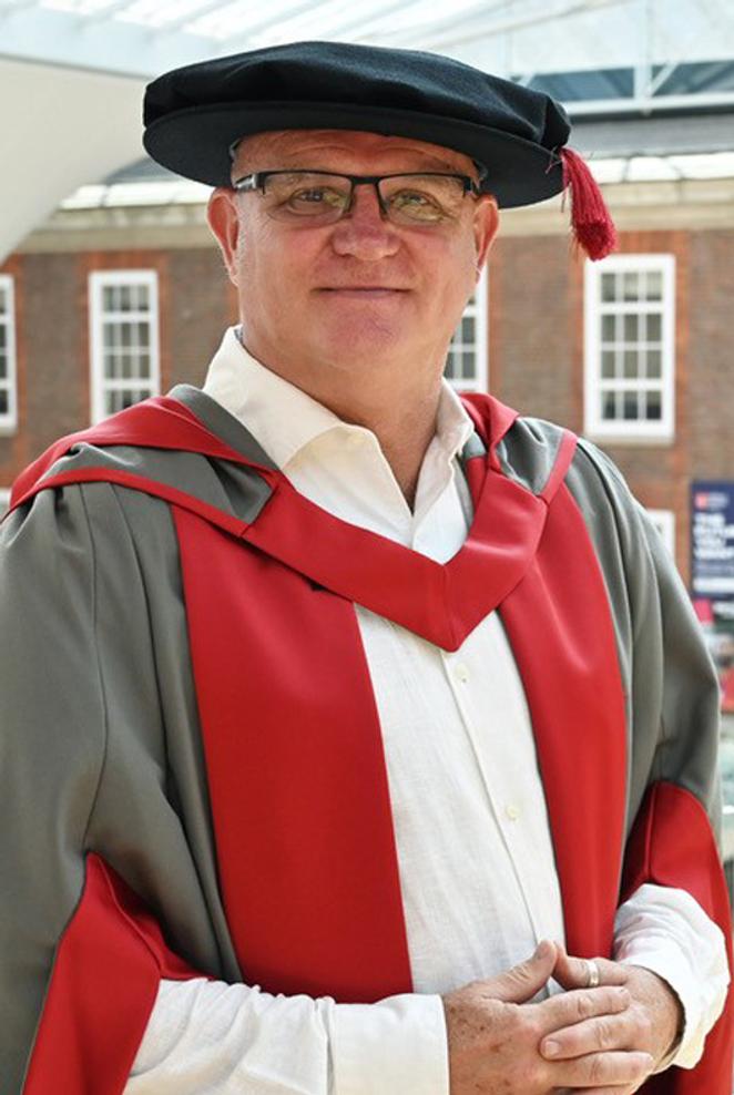 A man in honorary graduates cap and gown poses for a photo in a university building