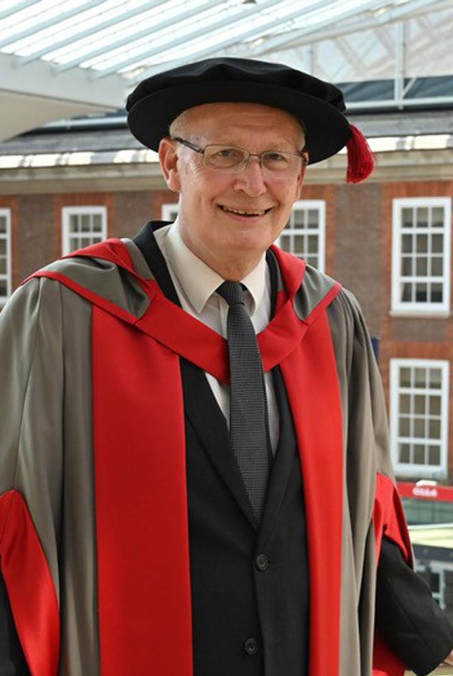 A man in honorary graduates cap and gown poses for a photo in a university building