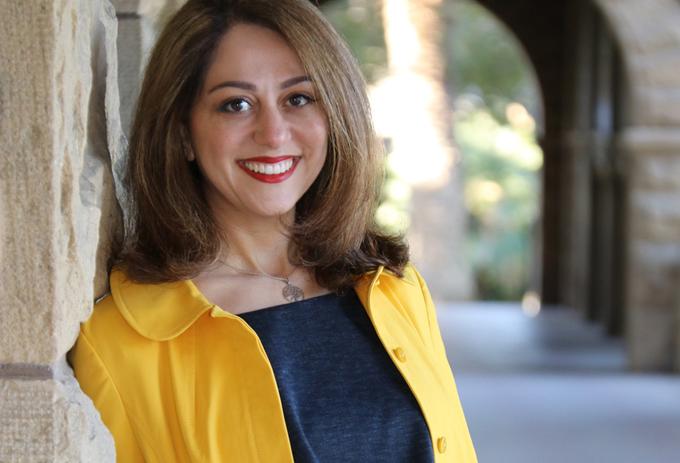 A portrait of Dr. Sara Nasserzadeh in a yellow coat in front of an old building with an archway