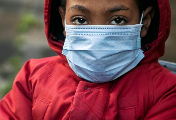 A photograph which is part of an art exhibition of a young black person with a blue surgical facemask on, wearing a red anorak with a hood, staring at the viewer