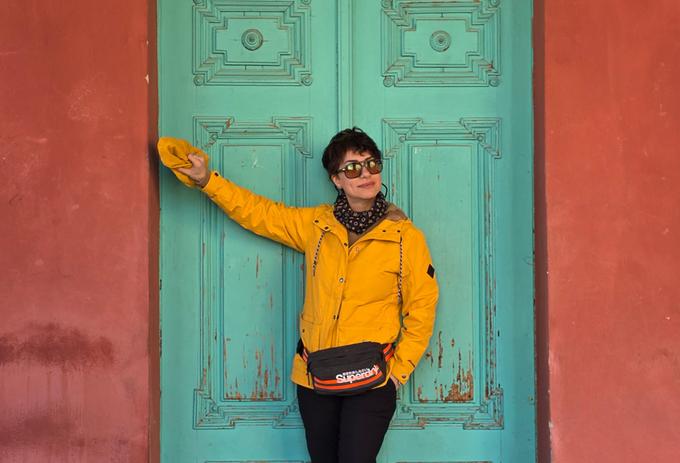A woman wearing a yellow jacket and sunglasses poses in front of a jade green door