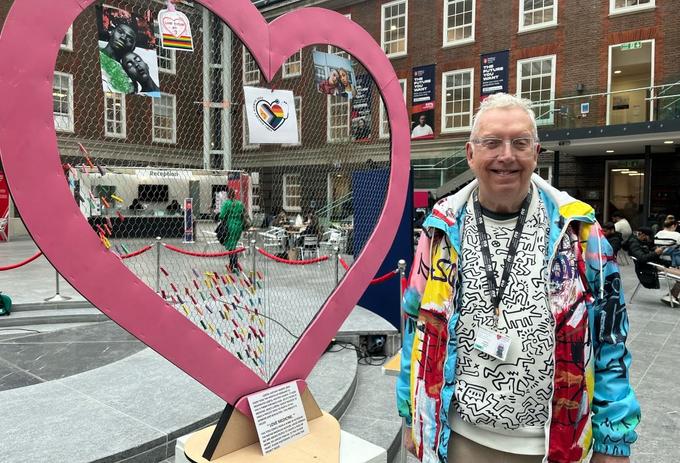 An older white man with white hair in a bright coloured graffiti-style jacket stands by a large cut-out sculpture of a heart