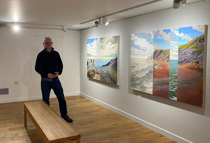 A portrait of Dr John Timberlake standing next to his paintings in the Granary Gallery, Berwick-Upon-Tweed
