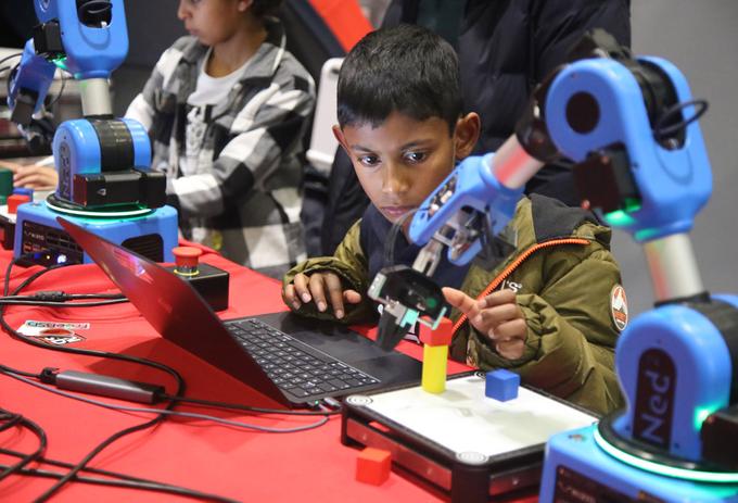 A young dark-haired boy sits at a laptop computer peering at a blue robot arm which is programming through the laptop keyboard