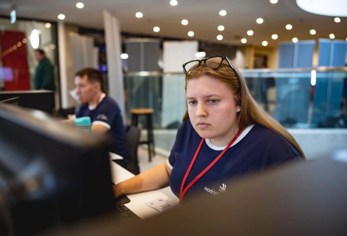 A young woman in a WorldSkills top and a lanyard sits at a computer as she takes part in a Digital Construction skills competition