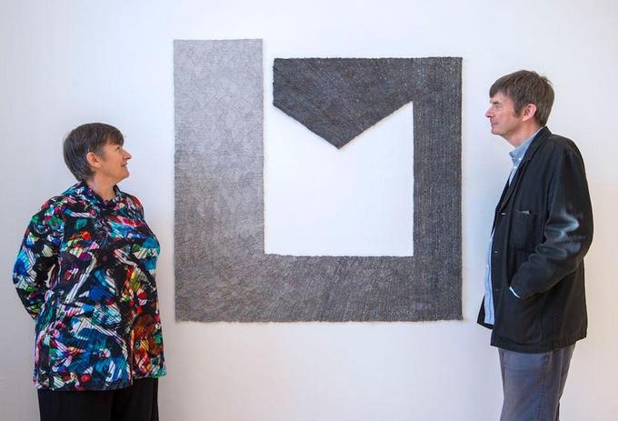 A brown-haired woman in a colourful top, and a brown-haired man in a jacket and blue shirt stand either side of a grey and black tapestry artwork looking closely at it.