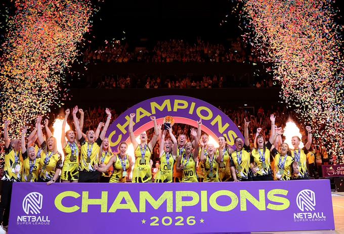 A netball team celebrates behind a sign saying 'champions' and a firework explosion a each side