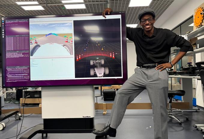 A man in a cap and glasses stands leaning against a large computer monitor, looking at the camera