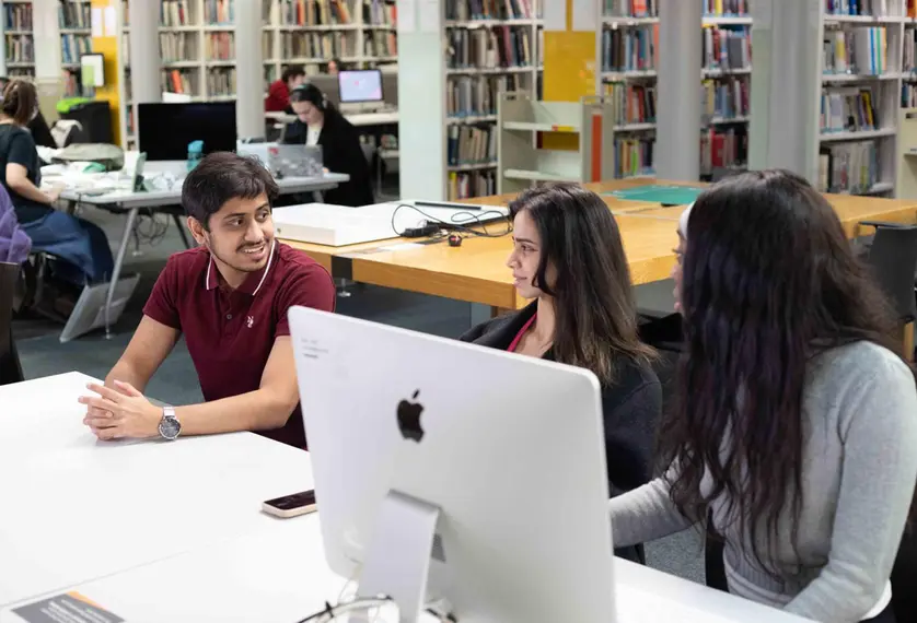 A group of students sit chatting in a work area with a desktop computer