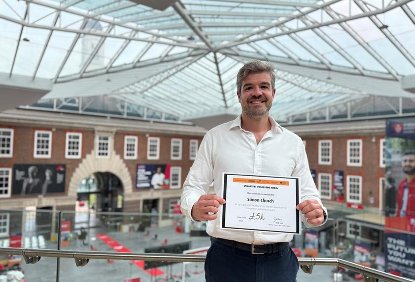 A MDX MBA student in a white shirt stands holding a certificate on the second floor balcony of the Quad