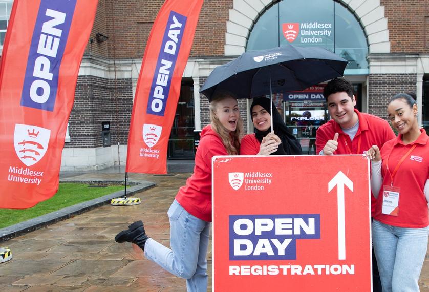 Four smiling students holding an umbrella, behind a sign pointing forwards to open day registration