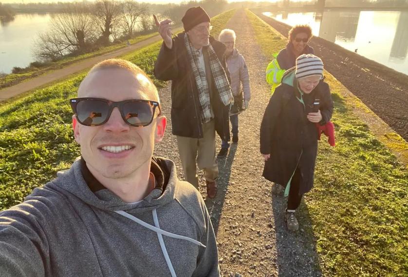 A young man in a hoodie and sunglasses takes a selfie in front of a group of older people in hats and scarves. They seem to be in the middle of a walk