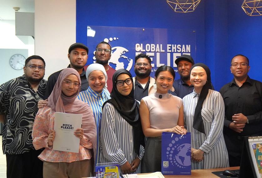 A group of men and women at Muslim anti-poverty charity Global Ehsan Relief stand in a group against a blue wall with their charity logo on it, smiling at the gamera