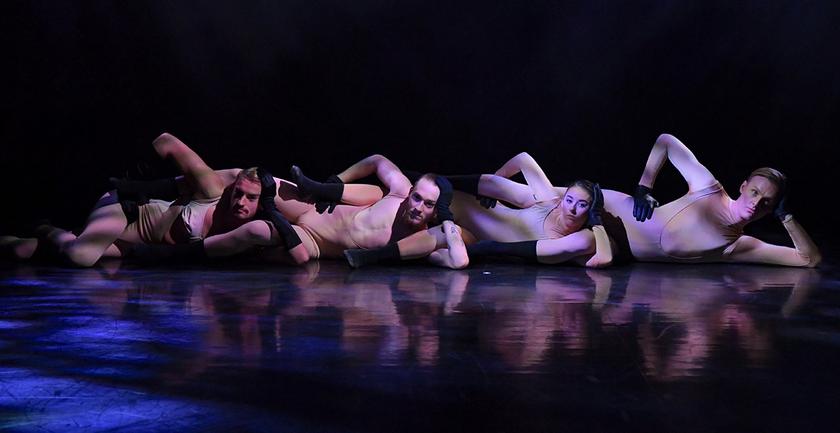 Four dancers mid-dance, lying on floor in a line with legs threaded through arms