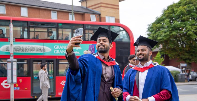 Two smiling male graduates in graduation robes taking a selfie outside College Building