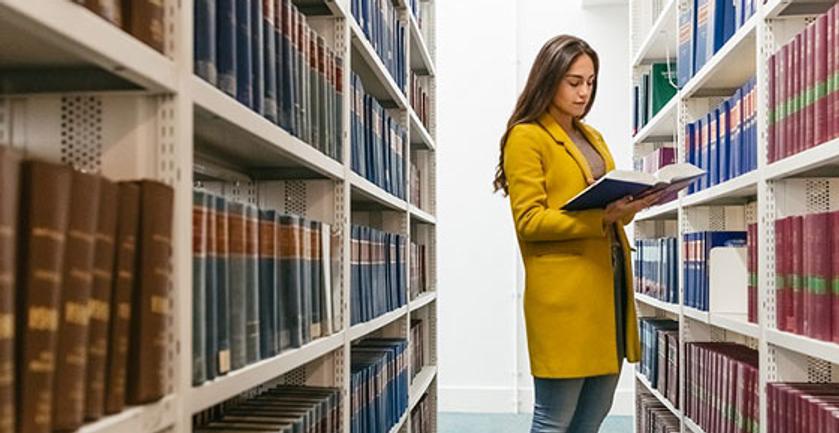 A woman in a yellow jacket standing in a library, surrounded by bookshelves filled with books.