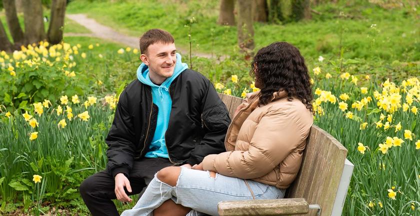Two students sit on bench with green behind them and daffodils