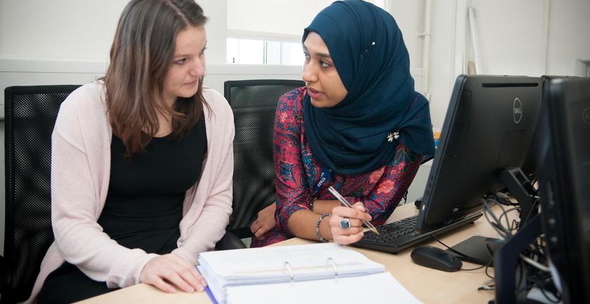 Two female students (one wearing a headscarf) sitting at a desk, talking
