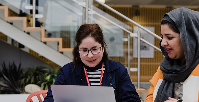 Two women sitting at a table with a laptop, discussing work projects and sharing ideas.