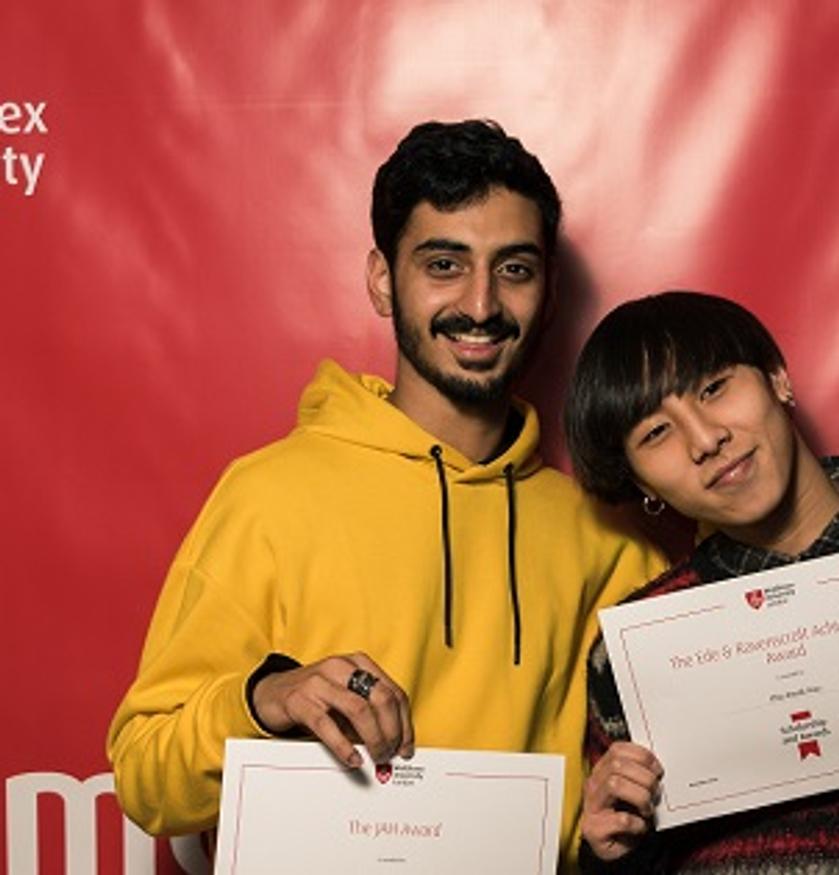 Two young men proudly displaying certificates in front of a red wall.