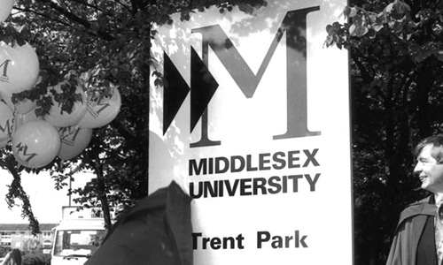 Two individuals in academic robes are unveiling a sign reading 'Middlesex University Trent Park' with several balloons next to it. © Middlesex University