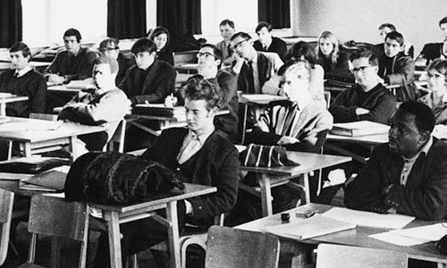A black and white photo of a class of students, male and female, sat at individual desks, listening to a barrister's lecture. Image credit: © Henry Grant Collection/London Museum