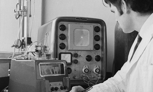 A black and white photo of a man wearing a white lab coat and taking notes at a desk while looking at a machine on the desk. Image credit: © Henry Grant Collection/London Museum