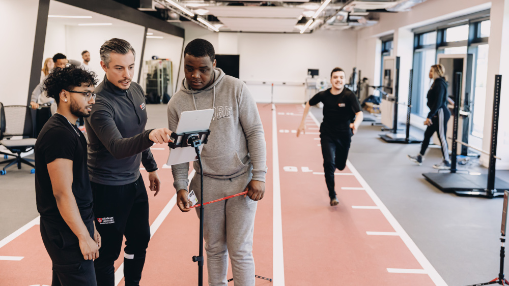 Three men in a gym huddled together, focusing on a tablet screen, engrossed in whatever they are viewing.