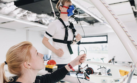 A male running on a large treadmill and wearing a breathing mask connected to cables with a woman next to the treadmill holding these cables.