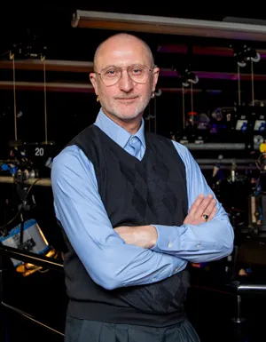 A man dressed in a sleeveless jumper and shirt stands with his arms folded in front of TV studio lights