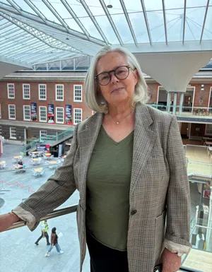 A woman wearing a jacket with grey hair and glasses standing in front of a building with lots of windows