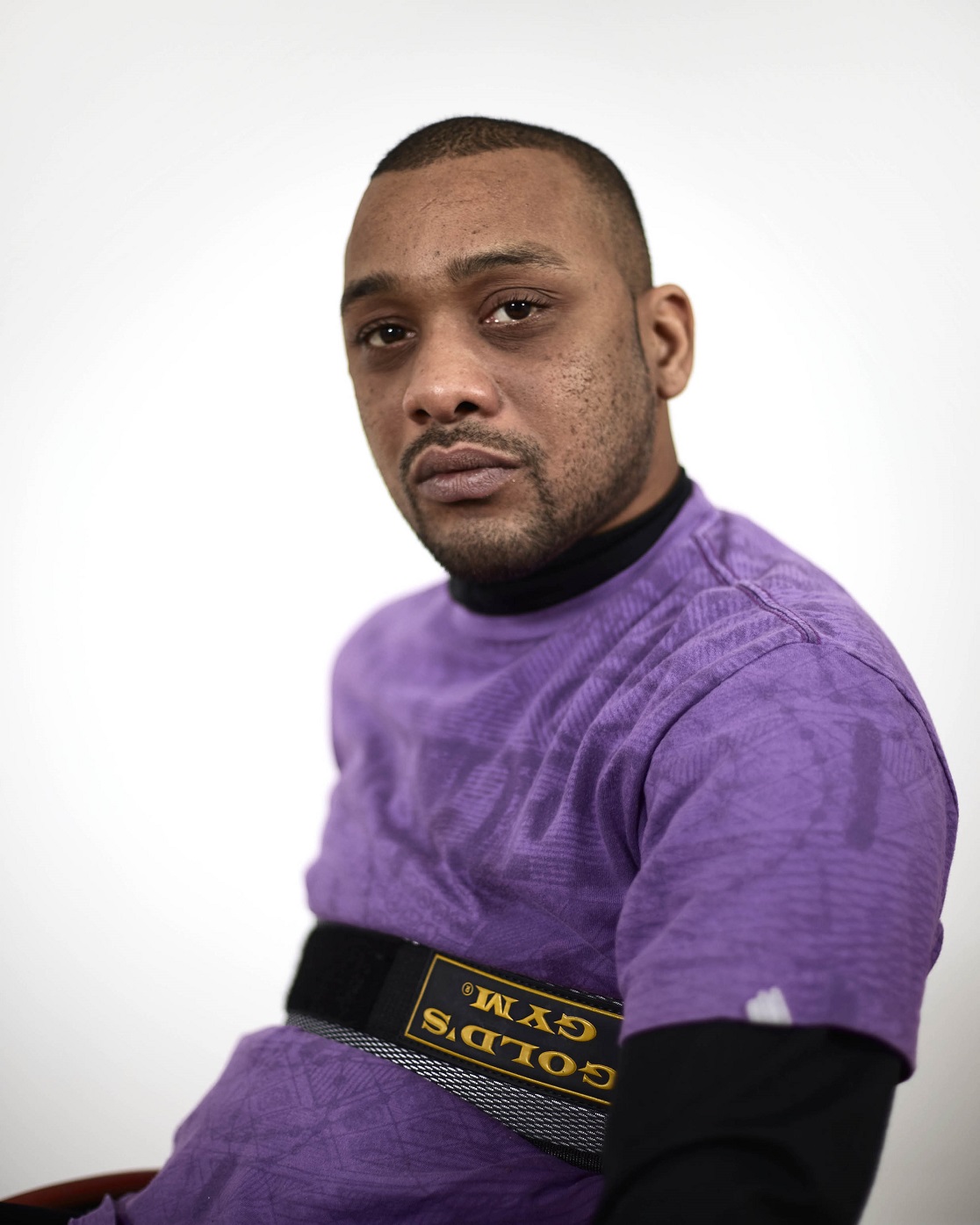 A young black man in a purple T-shirt, seated with his body turned to one side, looks directly at the viewer