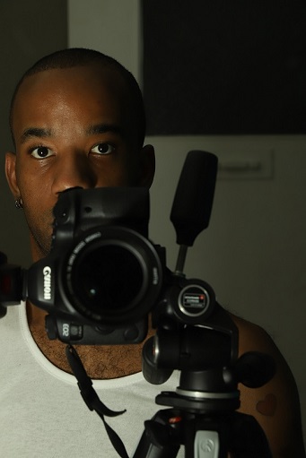 Dexter McLean, a young black man with close-cropped hair and wearing a white vest stands in front of a camera on a stand