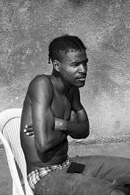 From Dexter McLean's series Olympic Gardens, a young black man, topless and with his arms crossed sits turned at an angle to the viewer, looking disconsolate